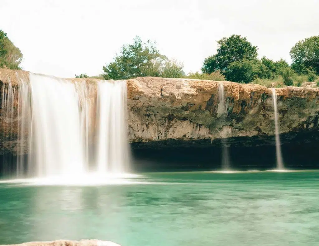 Cascade waterfall tumbling from a rocky overhang into a spacious pool, popular for swimming & picnics.